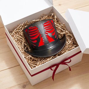 May include: A black top hat featuring a red ribcage cutout design, presented inside a white gift box filled with shredded paper filler. The box is tied with a thin red ribbon and sits on a light-colored wooden surface, showcasing the hat's structure.