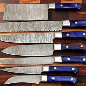 Op de afbeelding: Een set van zes keukenmessen met patronen stalen messen en blauwe houten handvatten. De handvatten zijn geaccentueerd met zilveren bolster en messing klinknagels. De messen zijn gerangschikt op een houten snijplank.