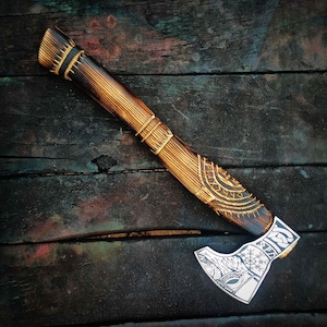 May include: A handcrafted axe with a wooden handle and a silver-colored blade. The handle is intricately carved with geometric patterns and wrapped with gold-colored bands. The blade features detailed engravings of Viking symbols.