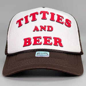 Puede incluir: Gorra de camionero marrón y blanca con las palabras "TITTIES AND BEER" en letras rojas. La gorra tiene una parte trasera de malla y una visera curva. El texto está centrado en el panel frontal.