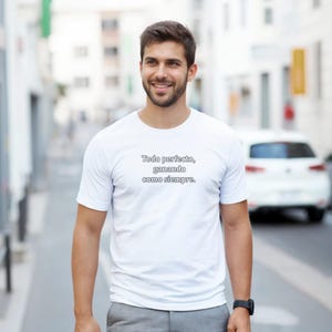 Könnte beinhalten: Weißes T-Shirt mit dem Text "Todo perfecto, ganando como siempre" in Schwarz. Das T-Shirt wird von einer Person im Freien getragen. Die Person lächelt und trägt eine schwarze Uhr.