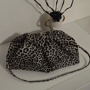 Op de afbeelding: Een zachte, geplooide clutch tas met een zwart-wit luipaardprint. De tas heeft een afgeronde, volumineuze vorm met een bijpassende dunne schouderband aan de zijkanten, wat zorgt voor een geplooid effect langs de opening aan de bovenzijde.