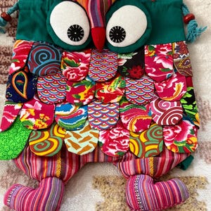Puede incluir: Una mochila de cordón con forma de búho, con una parte superior de tela verde, ojos blancos grandes y pupilas negras. El cuerpo está cubierto de parches de tela superpuestos con varios patrones florales, geométricos y de cachemira coloridos. Dos piernas de tela a rayas sobresalen de la parte inferior.