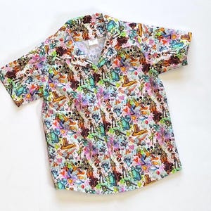 May include: Short-sleeved button-down shirt with a colorful pattern featuring various cartoon characters. The shirt has a white background with a collage of characters in shades of blue, green, pink, and brown. The shirt has a classic collar and short sleeves.