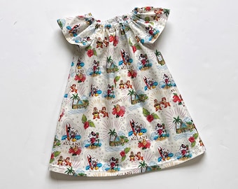May include: A white dress with a tropical print featuring Mickey and Minnie Mouse, palm trees, and surfboards. The dress has a gathered neckline and flutter sleeves. The word "Aulani" is printed on the dress.