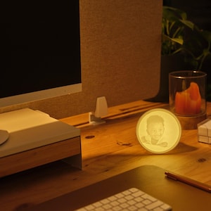 Puede incluir: Un retrato redondo e iluminado de una persona es el punto focal en un escritorio de madera. Una vela naranja encendida en un portavelas de vidrio, un monitor de computadora y un teclado también son visibles. La escena está cálidamente iluminada.