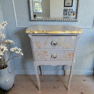 May include: A gray two-drawer side table with gold leaf trim and floral design. The table has crystal knobs and slender, curved legs. A mirror hangs above the table, reflecting a window and artwork.