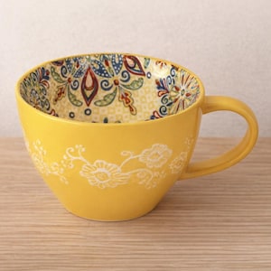 Peut inclure: Une tasse en céramique jaune avec un motif floral. La tasse a une anse et un motif floral décoratif à l'extérieur. L'intérieur de la tasse présente un motif coloré et complexe. La tasse est posée sur une surface en bois.