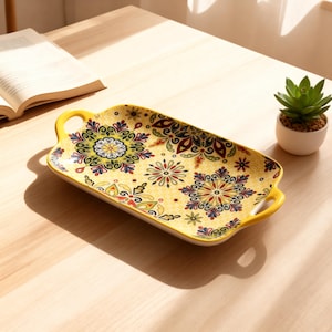 Può includere: Un vassoio rettangolare in ceramica gialla con un motivo floreale verde, rosso, blu e giallo. Il vassoio ha due manici gialli ed è posto su una superficie di legno. Accanto al vassoio c'è una piccola pianta grassa in un vaso bianco.