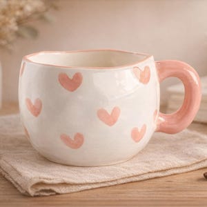 Peut inclure: Une tasse en céramique blanche avec une forme arrondie et une anse rose. La tasse est décorée de cœurs roses. La tasse est posée sur une serviette en lin beige sur une surface en bois. Des fleurs séchées sont en arrière-plan.