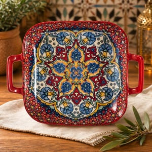 May include: Square ceramic serving dish with a vibrant floral pattern in red, blue, yellow, and white. The dish has a red border and two red handles. The design is reminiscent of traditional tilework.