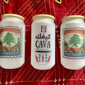 Puede incluir: Tres tarros de vidrio blanco con tapas de madera. Dos tarros presentan un diseño similar a un sello con un cedro libanés y la palabra "LEBANON". El tarro central tiene texto en árabe e inglés, junto con una ilustración de taza de café.
