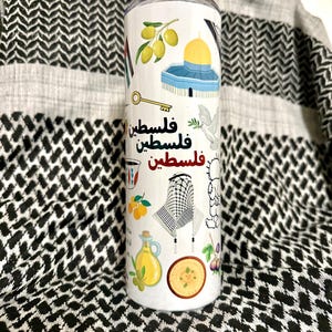 May include: A white travel mug decorated with Palestinian motifs, such as olives, a key, a dove, and the Dome of the Rock. The word "Palestine" is written in Arabic. The background is a black and white chequered pattern.