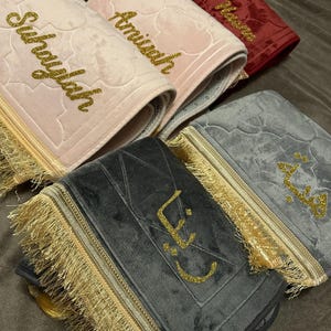 Könnte beinhalten: Eine Sammlung von Gebetsteppichen in verschiedenen Farben, darunter Rosa, Rot und Grau. Jeder Teppich hat goldglitzernde Buchstaben mit Namen und dekorative goldene Fransen. Die Teppiche sind gefaltet und gestapelt, was ihre weiche Samtstruktur zeigt.