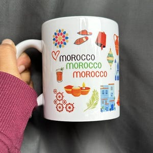 May include: White ceramic mug featuring colorful illustrations and the word "morocco" repeated in green and red. The mug is adorned with various Moroccan-themed icons, including a heart, a glass of tea, and traditional patterns.
