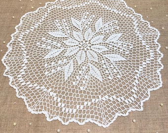 May include: A white, circular, crocheted doily featuring a floral centre design and a scalloped edge. The intricate lace pattern is visible throughout the doily, which is placed on a neutral-toned surface. Decorative pearls are scattered around the doily.