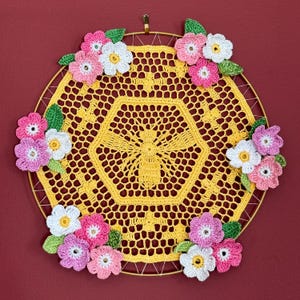 May include: A handmade, yellow crochet dream catcher with a bee design in the center. The dream catcher is adorned with pink, white, and green crochet flowers and leaves, and is suspended from a gold metal ring.