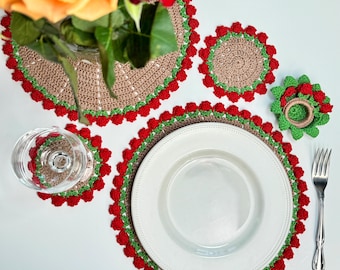 May include: A collection of handmade crochet placemats, coasters, and a napkin ring. The placemats and coasters are tan with green and red detailing. A white plate and glass are on the placemats. A silver fork is on the right.