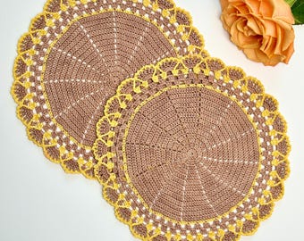 May include: Two handmade crochet doilies. The doilies are brown with a yellow scalloped edge. The doilies are circular with a radial pattern. A peach rose is in the upper right corner.