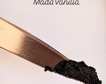May include: A silver-toned metal knife blade holds a small, textured mound of dark brown, granular vanilla bean paste. The text Mada Vanilla is printed in a black, cursive font above the knife blade against a plain, light-colored background.