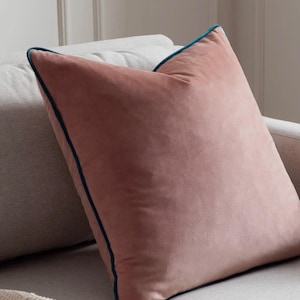 May include: A square cushion in a dusty rose hue, edged with teal blue piping. The cushion, made from a soft, velvety fabric, rests on a light grey sofa. A decorative item for home interiors.
