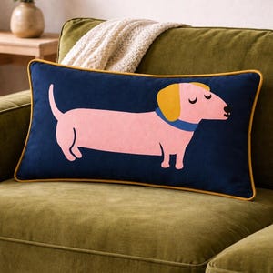 Peut inclure: Un coussin rectangulaire décoratif avec un motif de teckel rose sur fond bleu marine, bordé de jaune. Le chien a une oreille jaune et un collier bleu. Le coussin est posé sur un canapé vert.