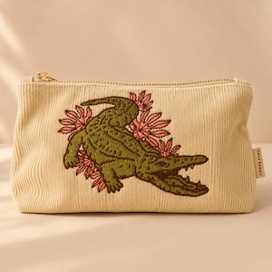 May include: A beige corduroy zippered pouch featuring an embroidered green alligator surrounded by pink floral shapes. A small fabric tag on the side of the pouch displays the text CASA RABBIT in black capital letters against a light background.