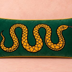 May include: A rectangular dark green velvet throw pillow featuring a large, yellow, winding snake graphic with black scale outlines and a red forked tongue. The pillow is finished with a vibrant yellow piped edge trim.