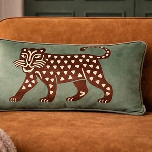 May include: A rectangular cushion with a teal background and a brown leopard design. The leopard has white spots and is walking to the left. The cushion is on a brown sofa.
