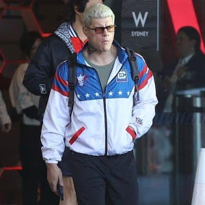 May include: A white, blue, and red zip-up jacket with a star pattern on the blue section. The jacket has a skull and logo patch on the chest and a matching patch on the sleeve. The person is wearing black pants and sunglasses.