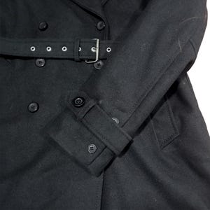 Puede incluir: Abrigo negro de doble botonadura con cintur&oacute;n y hebilla plateada. El abrigo presenta un cuello con solapas, pu&ntilde;os abotonados y varios botones negros. El abrigo est&aacute; hecho de una tela oscura y suave.
