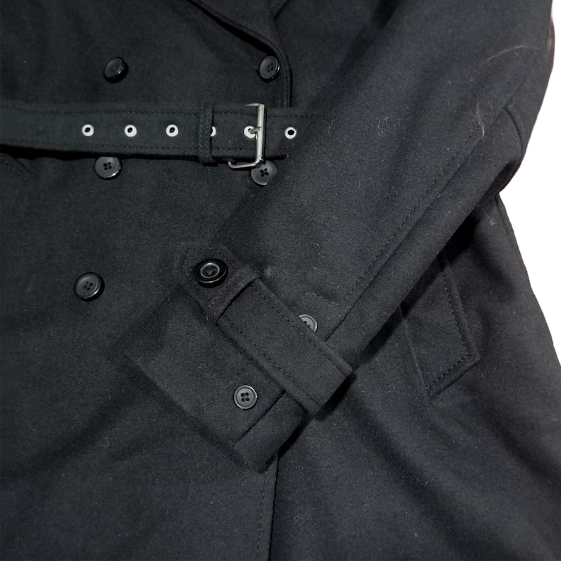 Puede incluir: Abrigo negro de doble botonadura con cintur&oacute;n y hebilla plateada. El abrigo presenta un cuello con solapas, pu&ntilde;os abotonados y varios botones negros. El abrigo est&aacute; hecho de una tela oscura y suave.