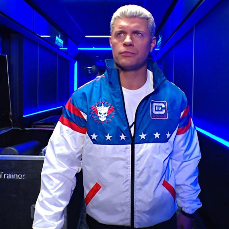 May include: A blue, white, and red zip-up jacket with a skull and crown emblem, five white stars, and a letter "R" patch. The jacket has a white body, blue shoulders, and red accents. The person is wearing a white t-shirt underneath.