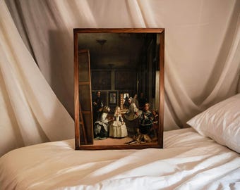 May include: A framed print of a classic painting, featuring a group of figures in a room with artwork on the walls. The frame is a dark wood, and the print is resting on a white bed with a white pillow. The background is a white sheet.