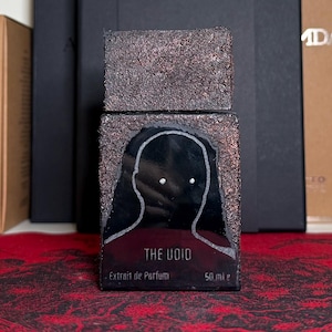 May include: A perfume bottle with a dark brown, textured exterior and a black front panel. The front features a white outline of a face with two white dots for eyes. The text "THE VOID" and "Extrait de Parfum" are visible. The bottle contains 50 ml.