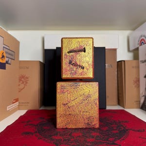 May include: Two-part gold-toned textured box with a nail and the word "Reliquia" on the top. The box is on a red patterned surface, with various boxes in the background.