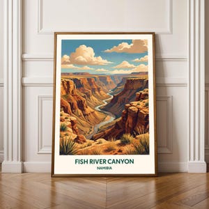 May include: A framed art print of Fish River Canyon in Namibia. The artwork depicts a scenic canyon landscape with a winding river, under a blue sky with fluffy white clouds. The color palette includes shades of brown, orange, and green.