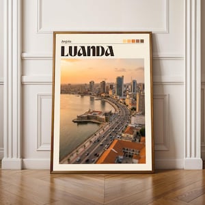May include: Framed print of Luanda, Angola, featuring a coastal cityscape at sunset. The print has a brown frame and includes the city name in bold, dark brown letters. The sky is a warm blend of orange and yellow.