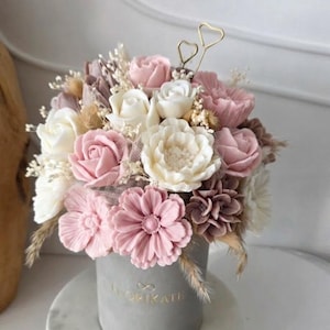 Pode incluir: Um arranjo floral em um recipiente cilíndrico cinza. O buquê apresenta flores rosa, brancas e malva, incluindo rosas e margaridas, com detalhes secos. Um fio dourado em forma de coração decora o topo. O recipiente tem a palavra "FLORIKATE" impressa.