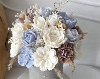Puede incluir: Arreglo floral en un recipiente cilíndrico gris. Las flores son una mezcla de blanco, azul claro y malva, con acentos de hierbas secas. El recipiente tiene la palabra "FLORIKATE" impresa. El arreglo está sobre una base de mármol.