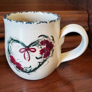 May include: Cream-colored ceramic mug with a heart-shaped floral design. The heart is outlined in green with red flowers and a red bow. The rim of the mug has a blue scalloped edge. The handle is a curved, off-white color.