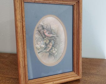 May include: A framed print featuring two birds perched on a flowering branch. The oval image is set against a blue background within a wooden frame. The artwork has a vintage aesthetic.