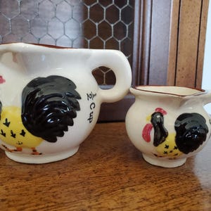 May include: Two ceramic cream pitchers with rooster designs. The larger pitcher has a capacity of 1/2 cup. The roosters are depicted with black, red, and yellow details. Both pitchers have a white base and brown trim.