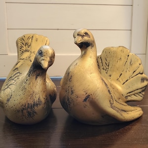May include: Two decorative bird figurines. The birds are painted gold with black accents, and are positioned on a dark brown surface. The bird on the left is smaller and has its tail feathers folded. The bird on the right has its tail feathers spread.