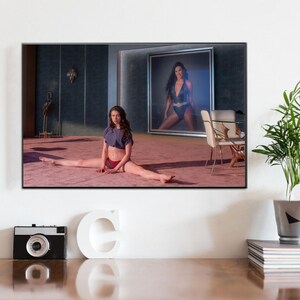 Peut inclure: Une impression d'art encadrée montrant une personne faisant le grand écart sur un tapis rose, portant un haut court violet foncé et un bas rouge. En arrière-plan, un grand portrait encadré montre une personne en maillot de bain bleu foncé.