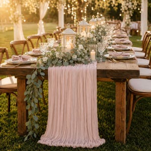 May include: A long wooden dining table set for an outdoor event, featuring a draped dusty pink fabric runner, white floral centerpieces with eucalyptus greenery, white lanterns, pillar candles, clear glassware, and folded pink cloth napkins.
