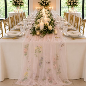 Puede incluir: Un camino de mesa de tul rosa pálido con delicados bordados de flores y mariposas, extendido sobre una larga mesa de comedor decorada con platos blancos, cubiertos dorados, cristalería y velas blancas en soportes de vidrio.