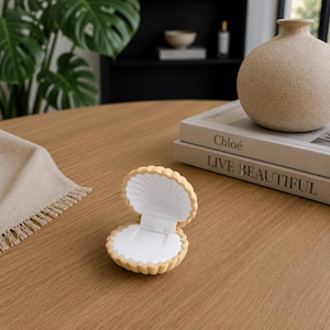 May include: A small, open jewelry box shaped like a seashell with a textured, light beige exterior and a smooth, white interior. The hinged container features a scalloped edge design and a flat white base inside, designed to hold a single piece of jewelry.
