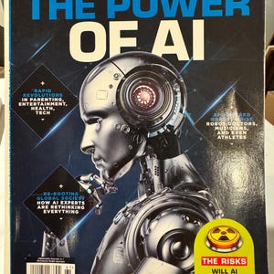 May include: The cover of TheStreet magazine features a metallic robot with the headline "THE POWER OF AI." Additional text includes "BREAKTHROUGHS CHANGING THE WORLD" and discussions on AI's impact on various sectors. A yellow sticker highlights "THE RISKS."