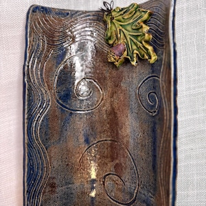 May include: Rectangular ceramic dish with a textured surface and a decorative leaf accent. The dish features a blue and brown color scheme with wave and swirl patterns. The leaf accent has green and purple tones.
